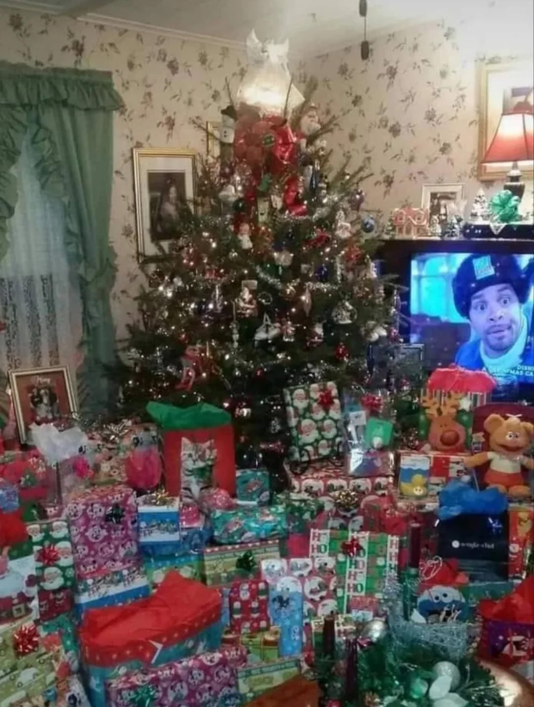 A living room with a decorated Christmas tree surrounded by numerous wrapped presents. The room has floral wallpaper, green curtains, and a TV showing a man making a surprised face.