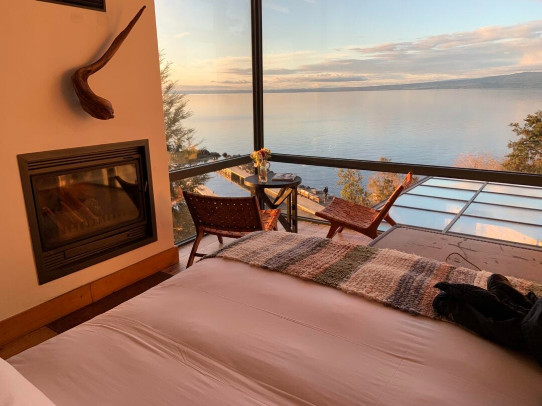 A cozy bedroom with a bed, fireplace, and woven chairs overlooks a large lake through floor-to-ceiling windows at sunset, with warm light filling the room and a scenic view outside.