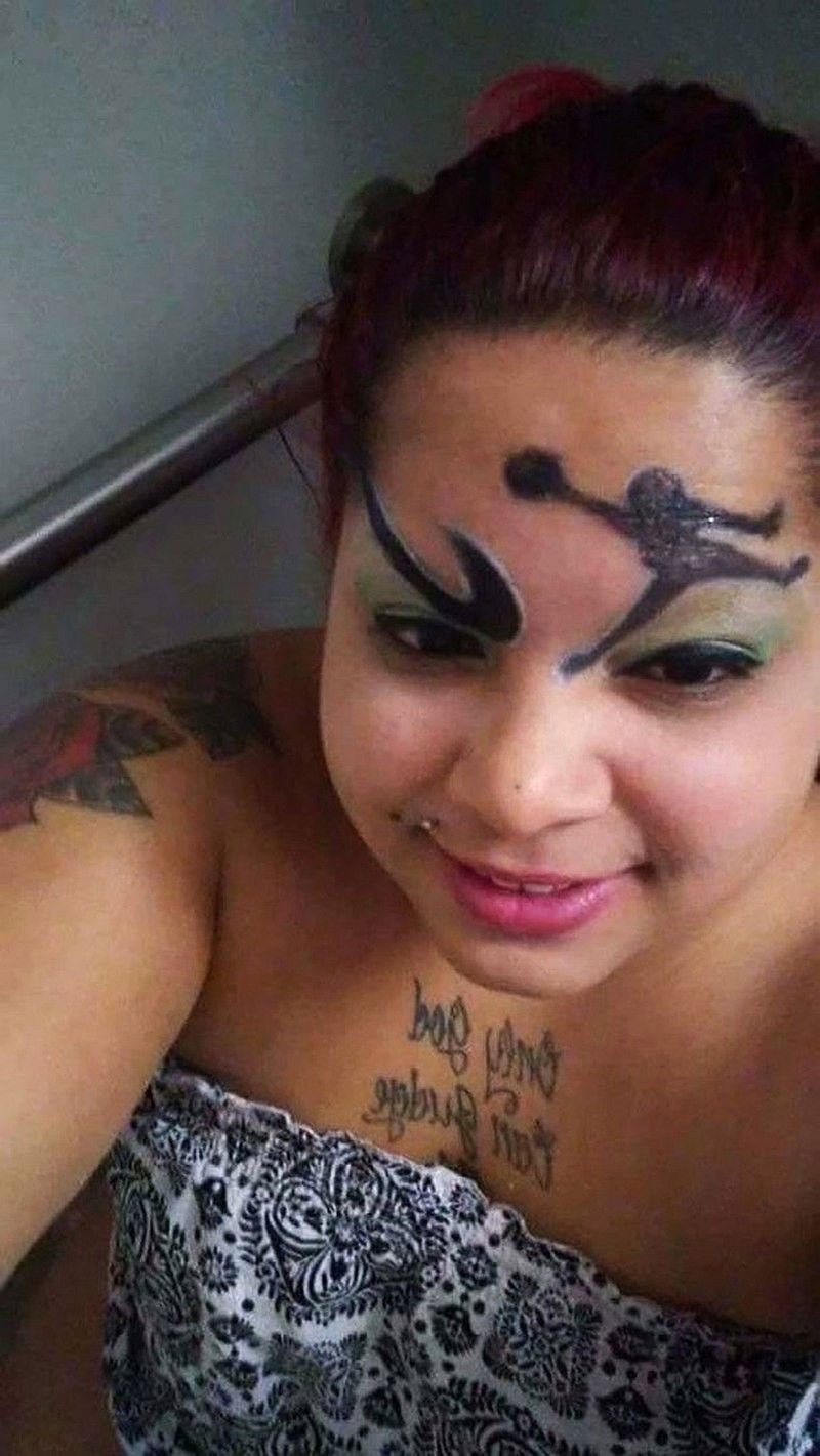 A woman with red hair smiles at the camera. She has a large tattoo of Tinker Bell on her forehead, green eyeshadow, a shoulder tattoo, and a chest tattoo that reads "Only God Can Judge." She is wearing a patterned top.