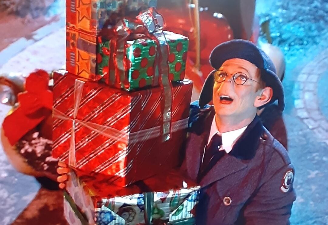 A person in a blue uniform and glasses smiles while holding a large stack of wrapped Christmas presents outdoors, with festive decorations and snow visible in the background.