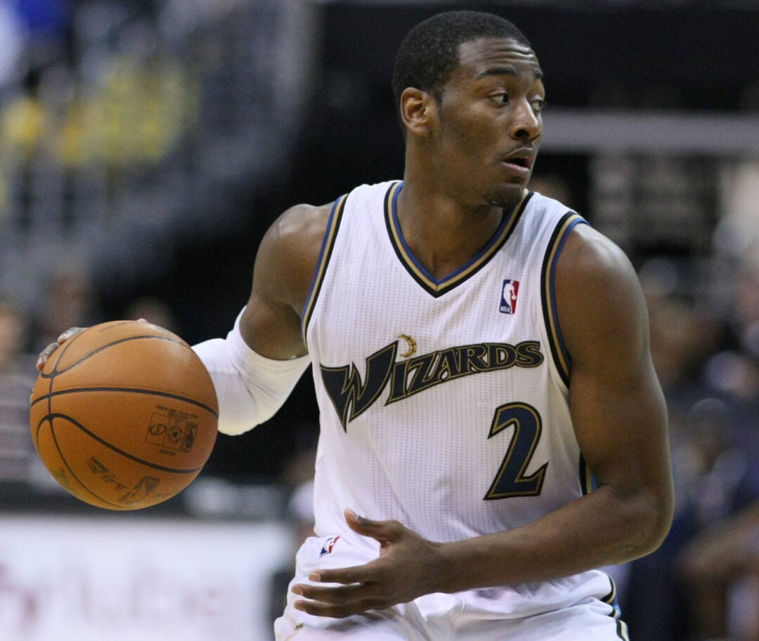 A basketball player in a white Wizards jersey with the number 2 dribbles a basketball during a game, looking to his left. The background shows a blurred crowd and court.