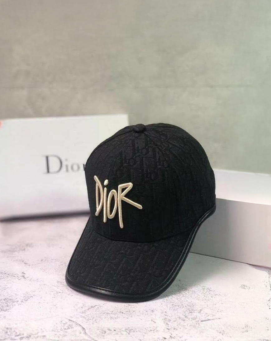 A black Dior baseball cap with the brand name "Dior" embroidered prominently in large, white letters on the front, displayed on a light surface with a white Dior box in the background.