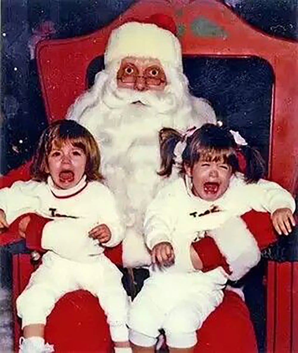 Two young children in white outfits sit on Santa's lap, both crying with distressed expressions. Santa is seated in a large red chair, looking straight at the camera with a serious face.
