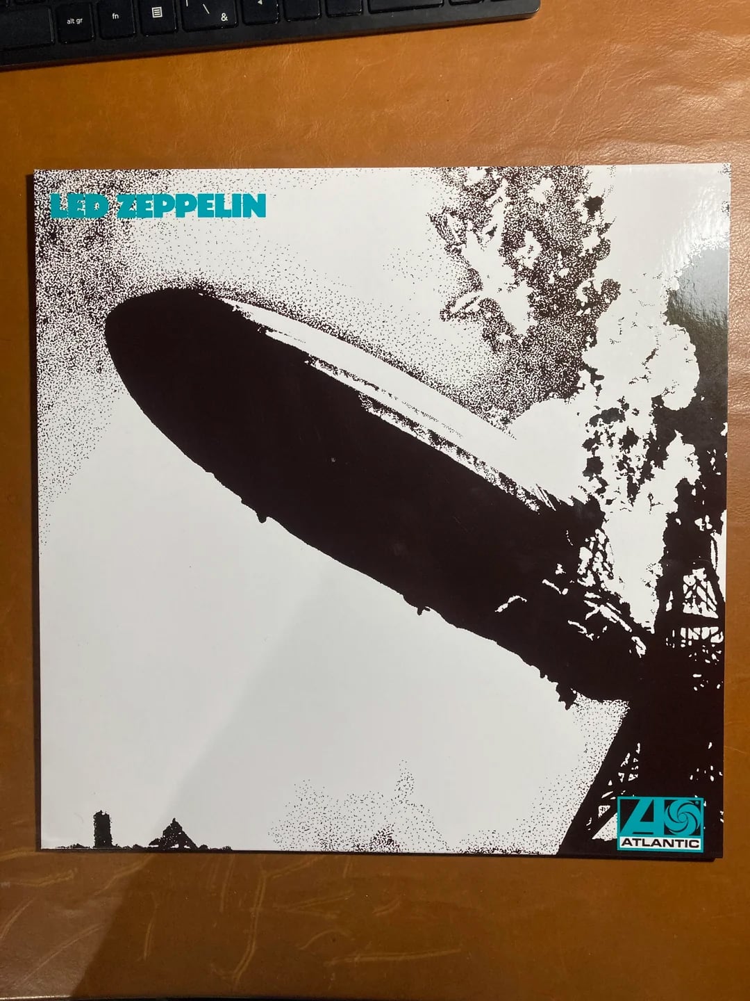 The image shows the iconic Led Zeppelin album cover, featuring a black-and-white photo of the Hindenburg airship disaster, with “Led Zeppelin” written in turquoise in the top left corner and the Atlantic Records logo in the bottom right.