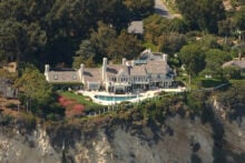 Aerial view of a large mansion with a pool, set atop coastal cliffs overlooking a sandy beach and the ocean, surrounded by trees and other homes. Waves gently reach the shore below.