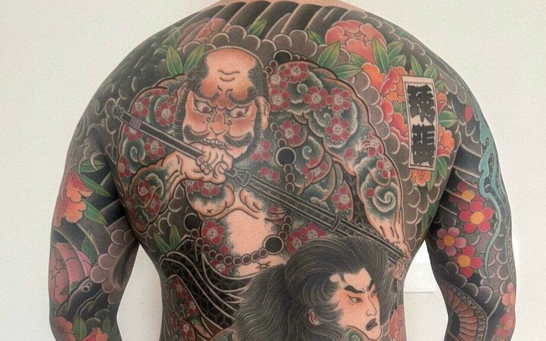 A person with a full-body traditional Japanese tattoo, featuring two large figures, elaborate patterns, flowers, and waves covering their back, arms, and legs, standing against a plain white background.