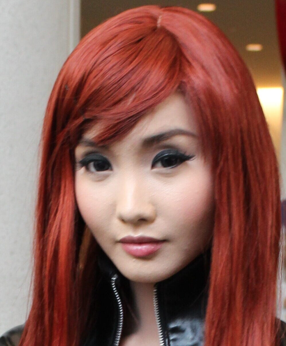A person with long, straight red hair and dramatic eye makeup is wearing a black outfit with a high collar, looking directly at the camera. The background is indoors with soft lighting.
