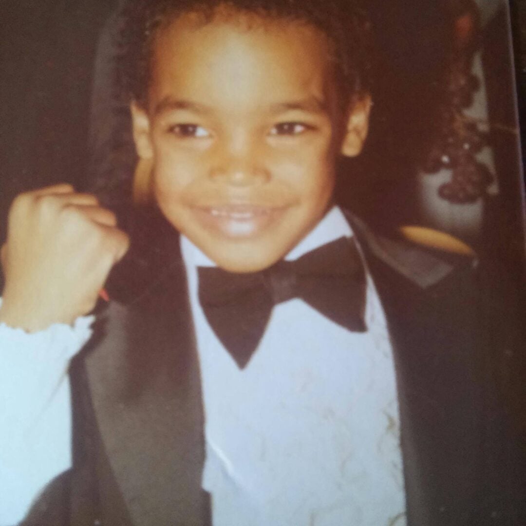 A young child smiles confidently at the camera, dressed in a black tuxedo with a bow tie and white ruffled shirt, holding up a fist in a celebratory gesture.