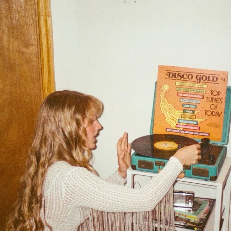 A woman with long, wavy hair plays a record on a teal turntable. The record cover behind her reads “Disco Gold: Top Tunes of Today.” She is wearing a fringed, white, textured blouse and appears to be placing the needle on the vinyl.