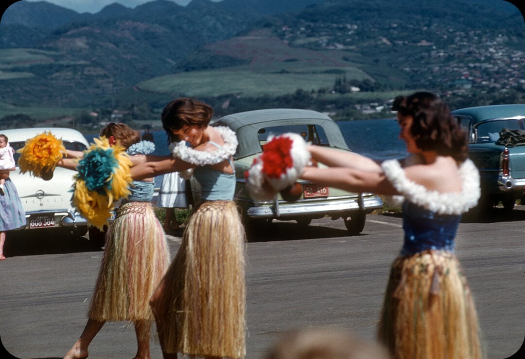 Three women in grass skirts and leis perform a hula dance with pom-poms outdoors, with vintage cars parked behind them and green mountains visible in the background.