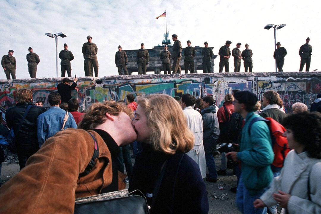 A couple kisses in front of the Berlin Wall, surrounded by a crowd. Soldiers stand on top of the graffiti-covered wall, with the Brandenburg Gate and a German flag visible in the background.