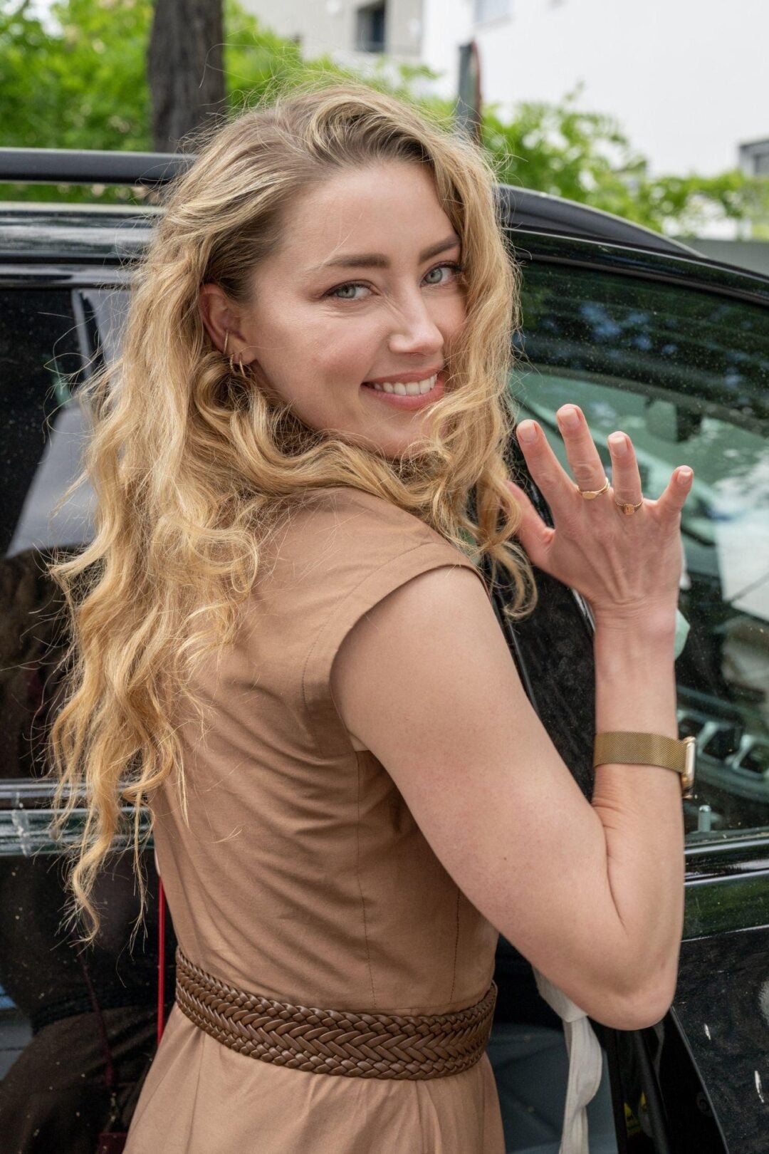 A woman with long, wavy blonde hair wearing a tan dress smiles and waves while standing next to a black car. She is looking over her shoulder at the camera.