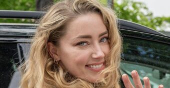 A woman with long, wavy blonde hair, wearing a tan dress and gold bracelet, smiles while turning toward the camera and waving, standing next to a black car outdoors.