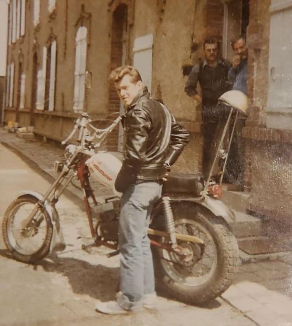 A man in a black leather jacket and jeans stands beside a vintage motorcycle on a street, looking at the camera. Two other men stand in a doorway behind him, partially visible. The buildings have brick walls and shuttered windows.