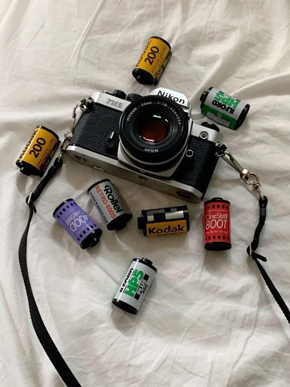 A Nikon film camera rests on a white fabric surface, surrounded by various 35mm film rolls including Kodak, Fuji, CineStill, and Ilford brands.