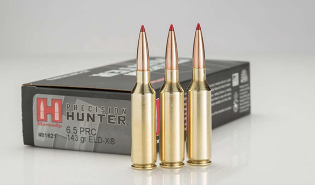 Three 6.5 PRC rifle cartridges with red-tipped bullets stand upright in front of a black and gray Hornady Precision Hunter ammunition box.