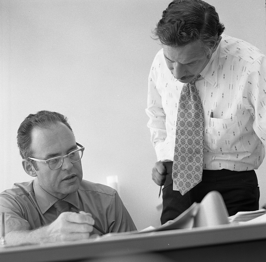 Two men in an office setting, one seated wearing glasses and looking at documents, while the other stands beside him, leaning forward and listening attentively. Both are dressed in business attire.