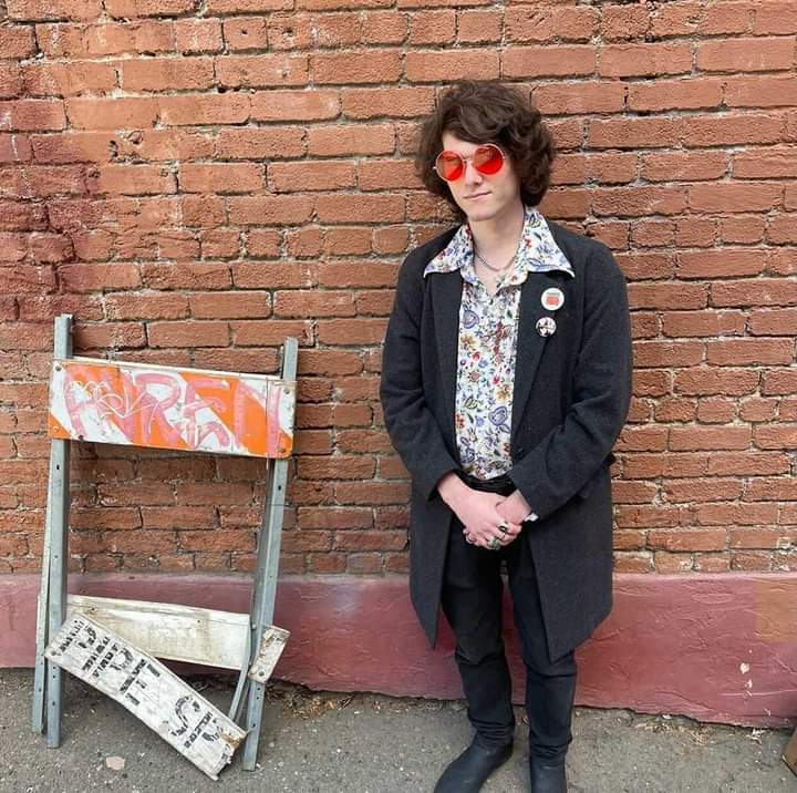 A person with curly hair, wearing red sunglasses, a floral shirt, and a dark coat stands against a brick wall next to a broken street barrier with graffiti.