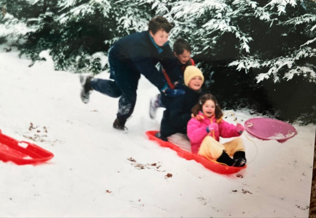 Four children play in the snow; two push a red sled holding two others. The child in front wears a pink coat and holds a purple sled. Snow-covered trees are in the background, and everyone appears to be having fun.