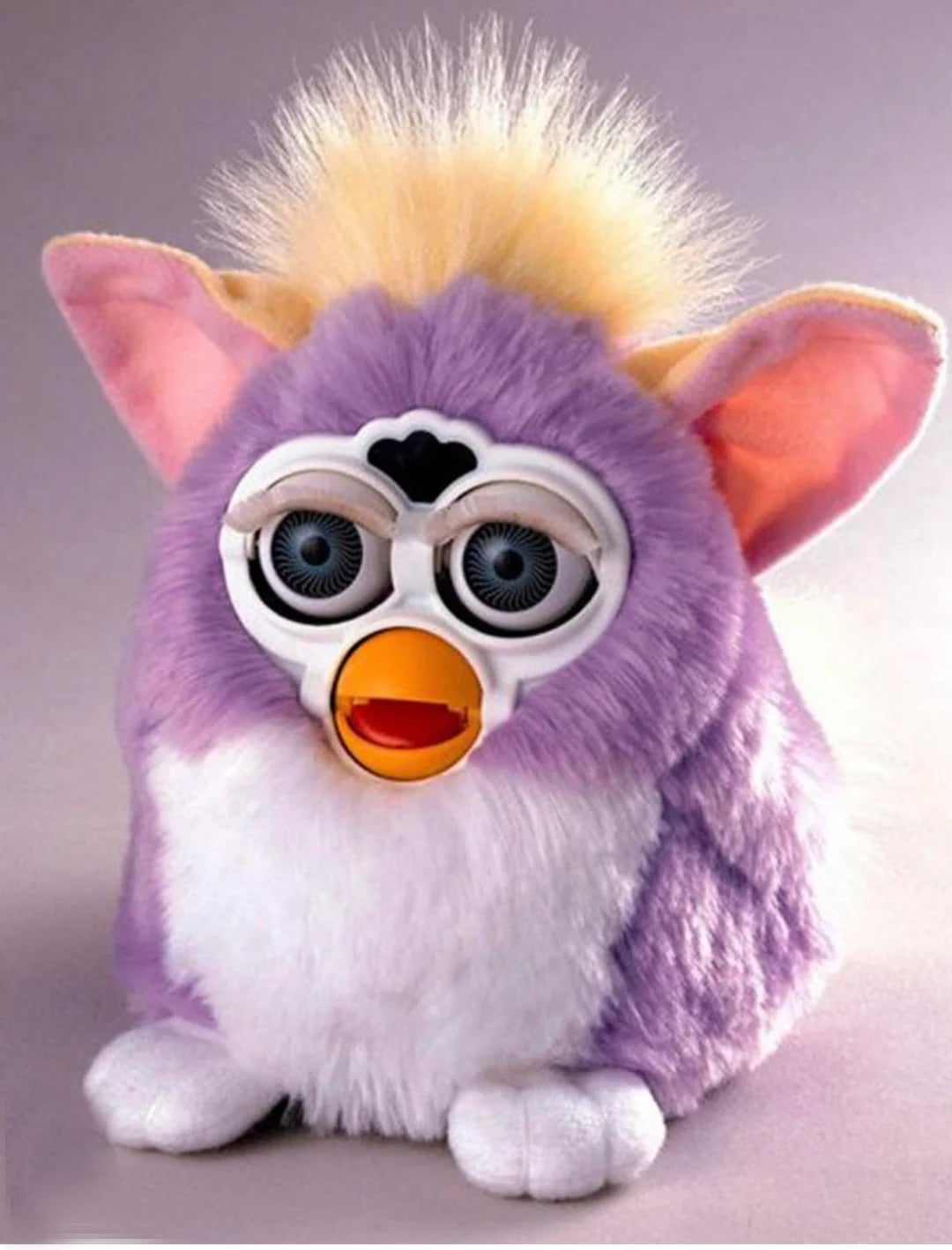 A purple and white Furby toy with big blue eyes, large pink ears, yellow tuft of hair, and an orange beak, sitting on a light surface against a neutral background.