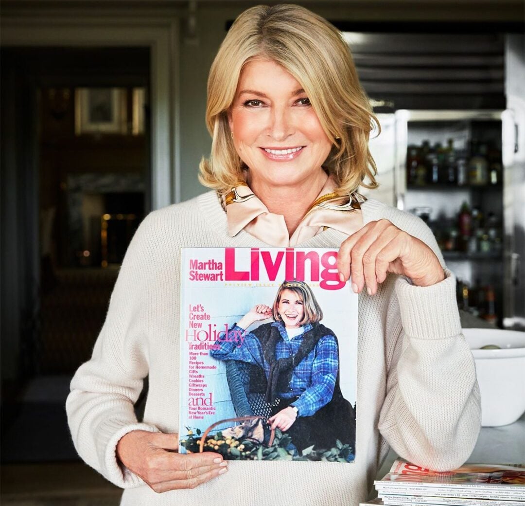 A smiling woman in a cream sweater stands indoors, holding a copy of Martha Stewart Living magazine featuring herself on the cover. A kitchen and stacked magazines are visible in the background.