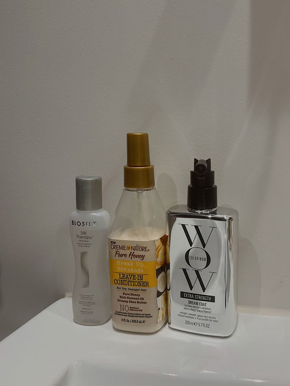 Three hair care products—Biosilk Silk Therapy, Creme of Nature Pure Honey Leave-In Conditioner, and Color Wow Extra Strength Dream Coat—are placed on a white bathroom sink against a plain white wall.