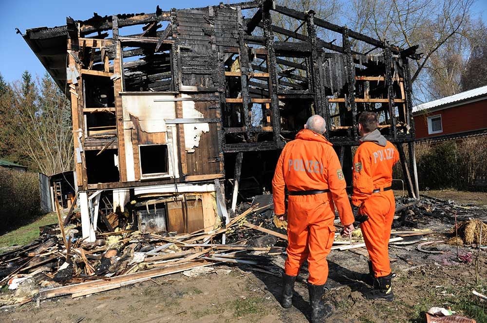 Two people in orange uniforms labeled "Polizei" stand in front of a severely burned and partially collapsed wooden building, surrounded by debris and remnants of the fire.