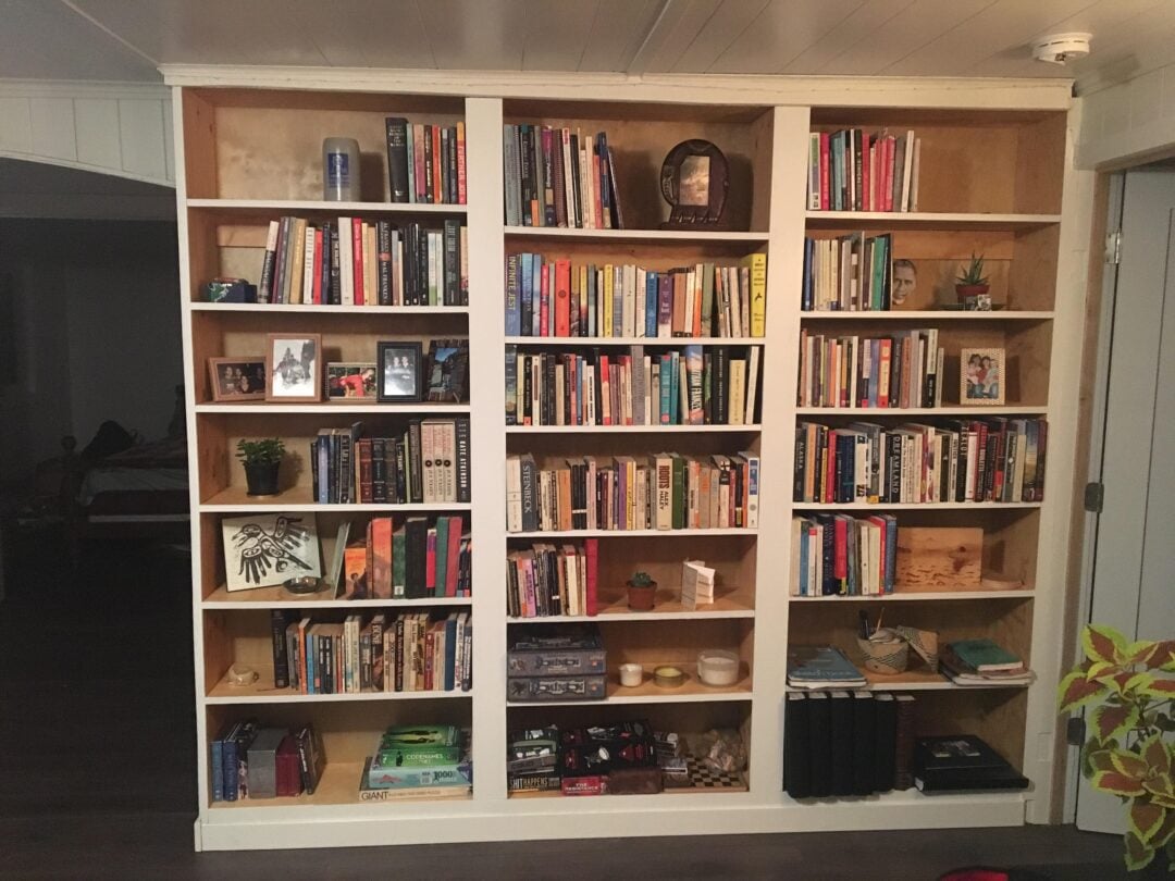A large white bookshelf filled with books, framed photos, small plants, decorative items, and a few board games, standing against a wall in a cozy room with dark floors and a white ceiling.
