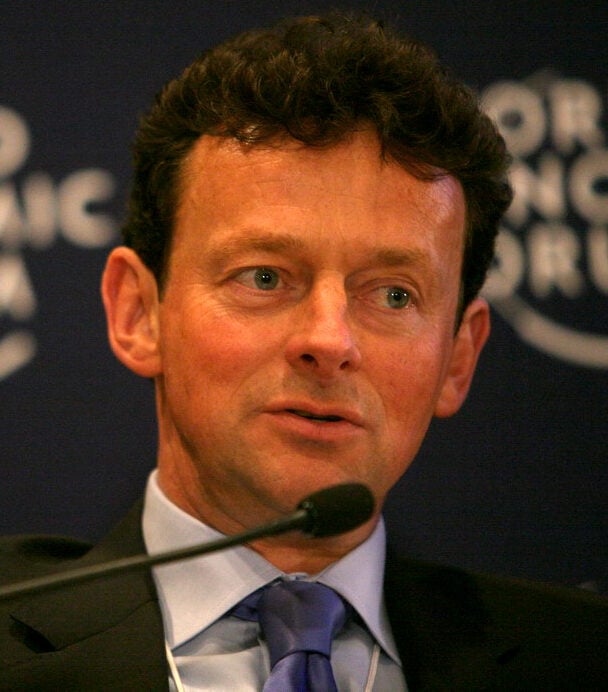 A man in a suit and tie speaks into a microphone against a dark blue backdrop with partially visible white text and logo, likely at an event or conference.