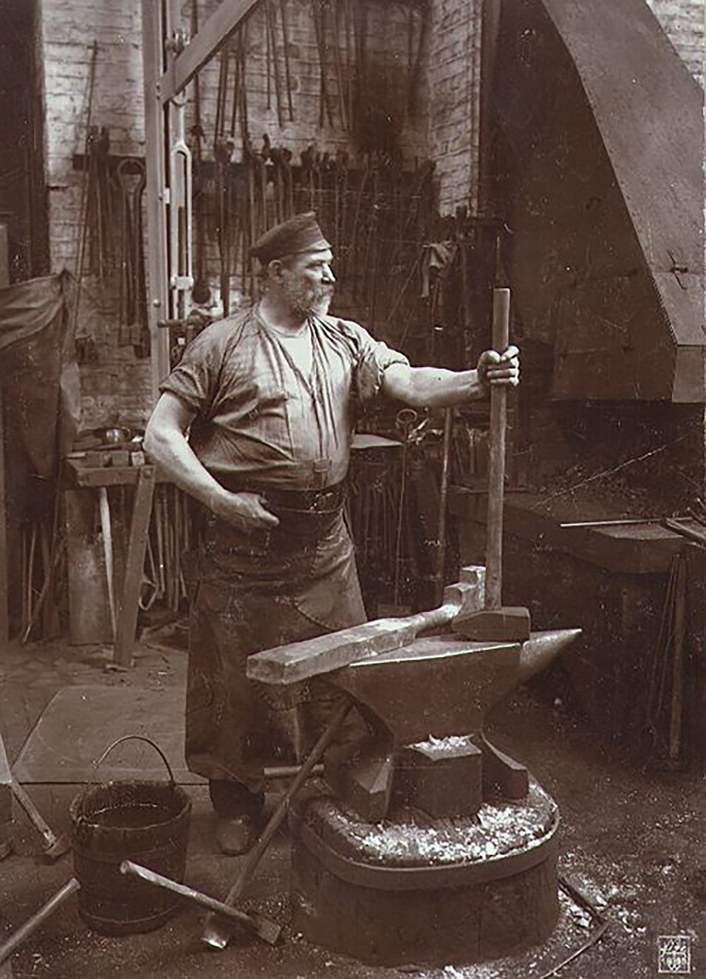 A black-and-white photo of a blacksmith standing in a workshop, holding a large tool with one hand and resting the other on his hip. An anvil, forge, and various tools are visible in the background.