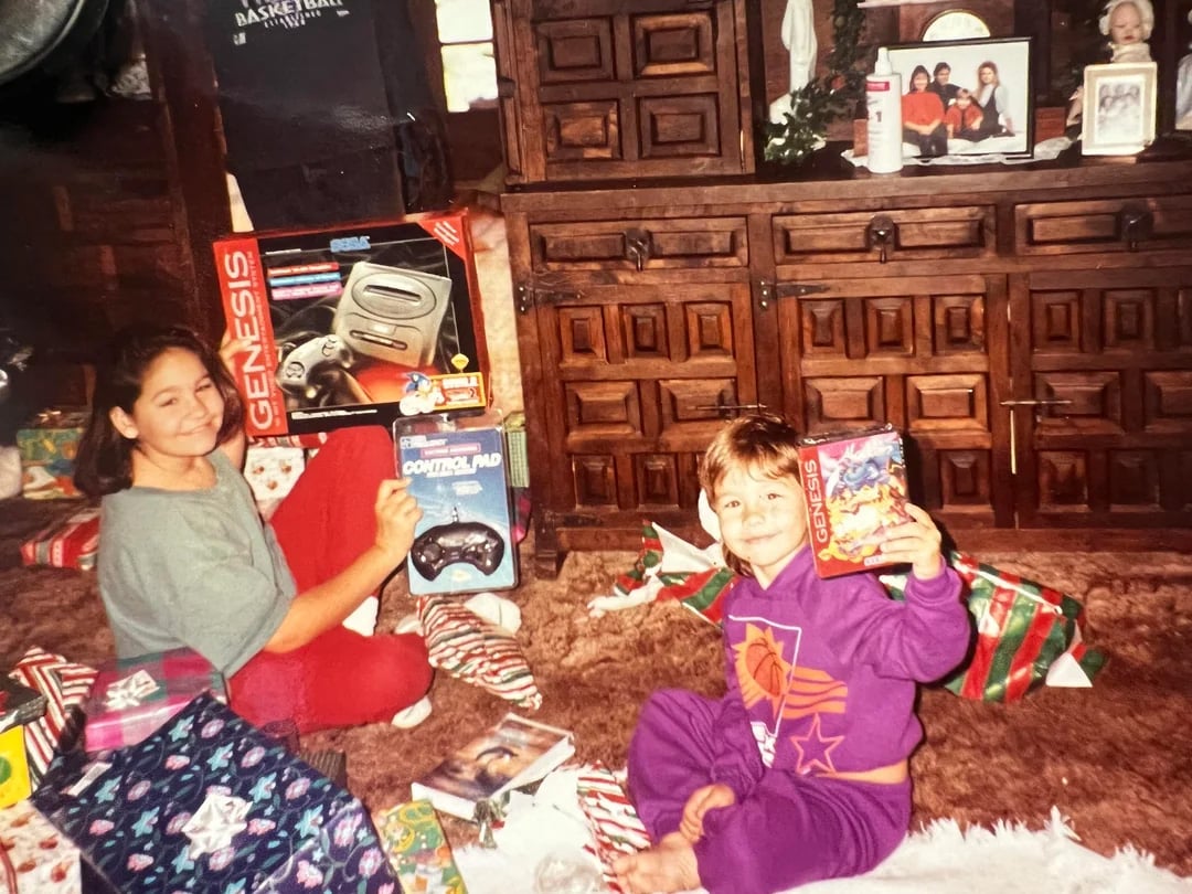 Two smiling children sit on a carpeted floor opening Christmas gifts. One holds a Sega Genesis console and controller, the other holds a game. Wrapping paper and presents are scattered around, with a wooden cabinet in the background.
