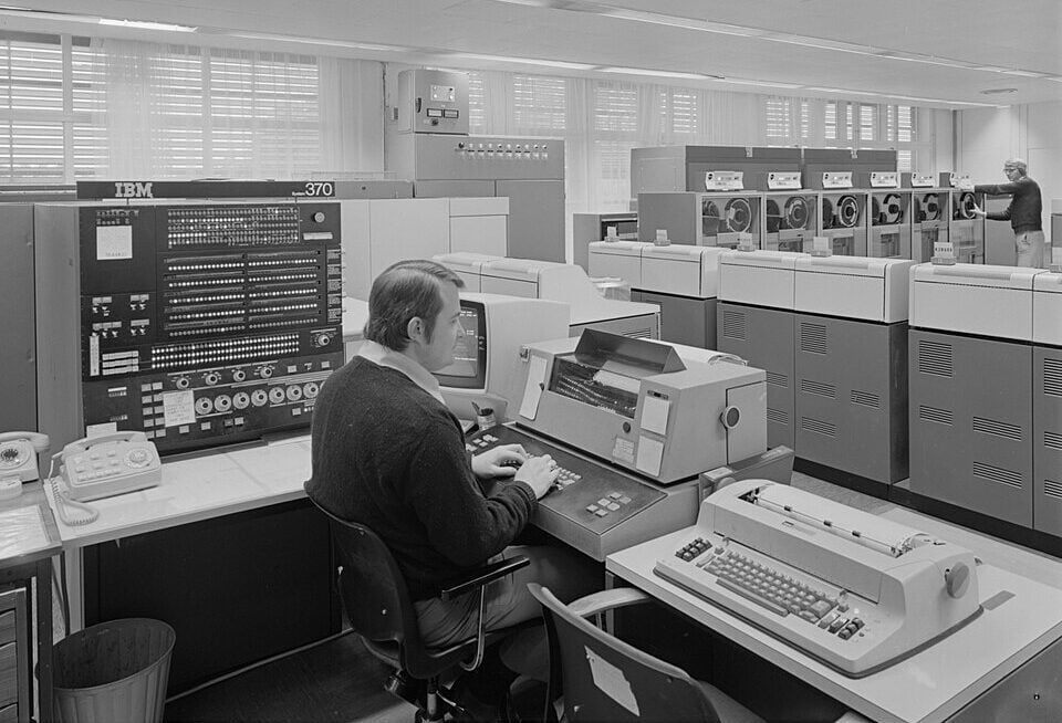 A man sits at a control desk operating an IBM computer system in a large room filled with mainframe computers and tape drives. Another person stands near computer equipment in the background. The scene is in black and white.