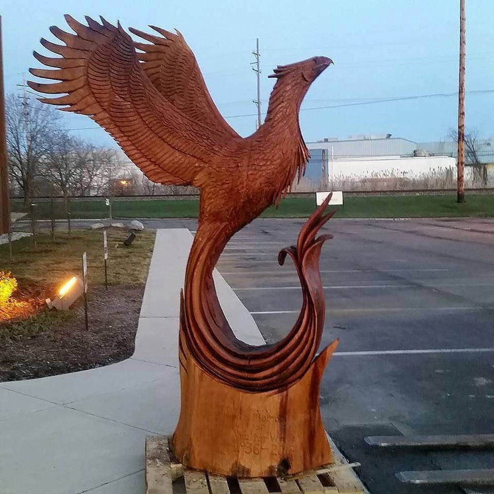 A large, detailed wooden sculpture of a phoenix with outstretched wings stands on a pallet near a parking lot at dusk. The carving highlights intricate feathers and a flowing tail.