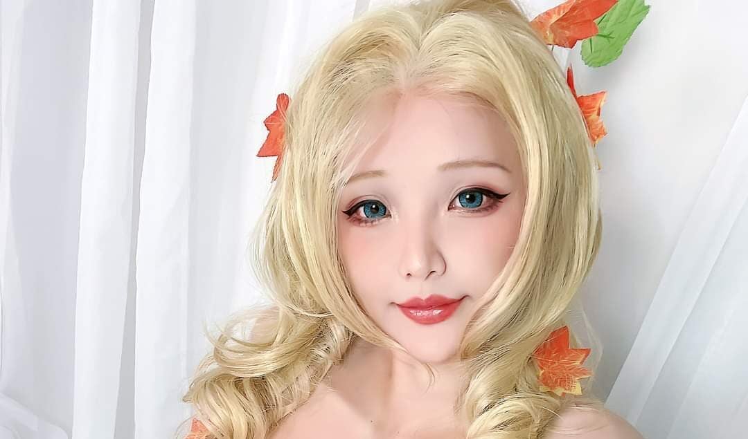 A woman with long blonde hair, styled with loose curls and leaves, poses in a costume adorned with green leaves covering her chest and lower body, sitting against white fabric.