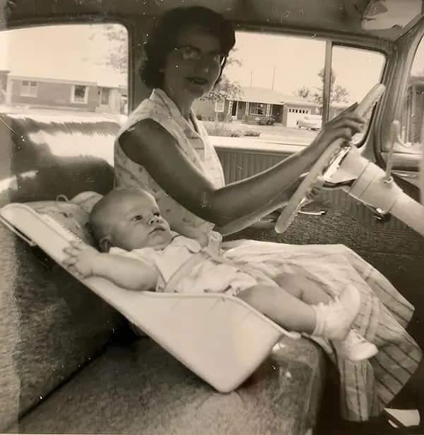 A woman drives a vintage car while a baby lies in a simple car seat on the front bench seat. The photo is black and white, with houses visible through the car window in the background.
