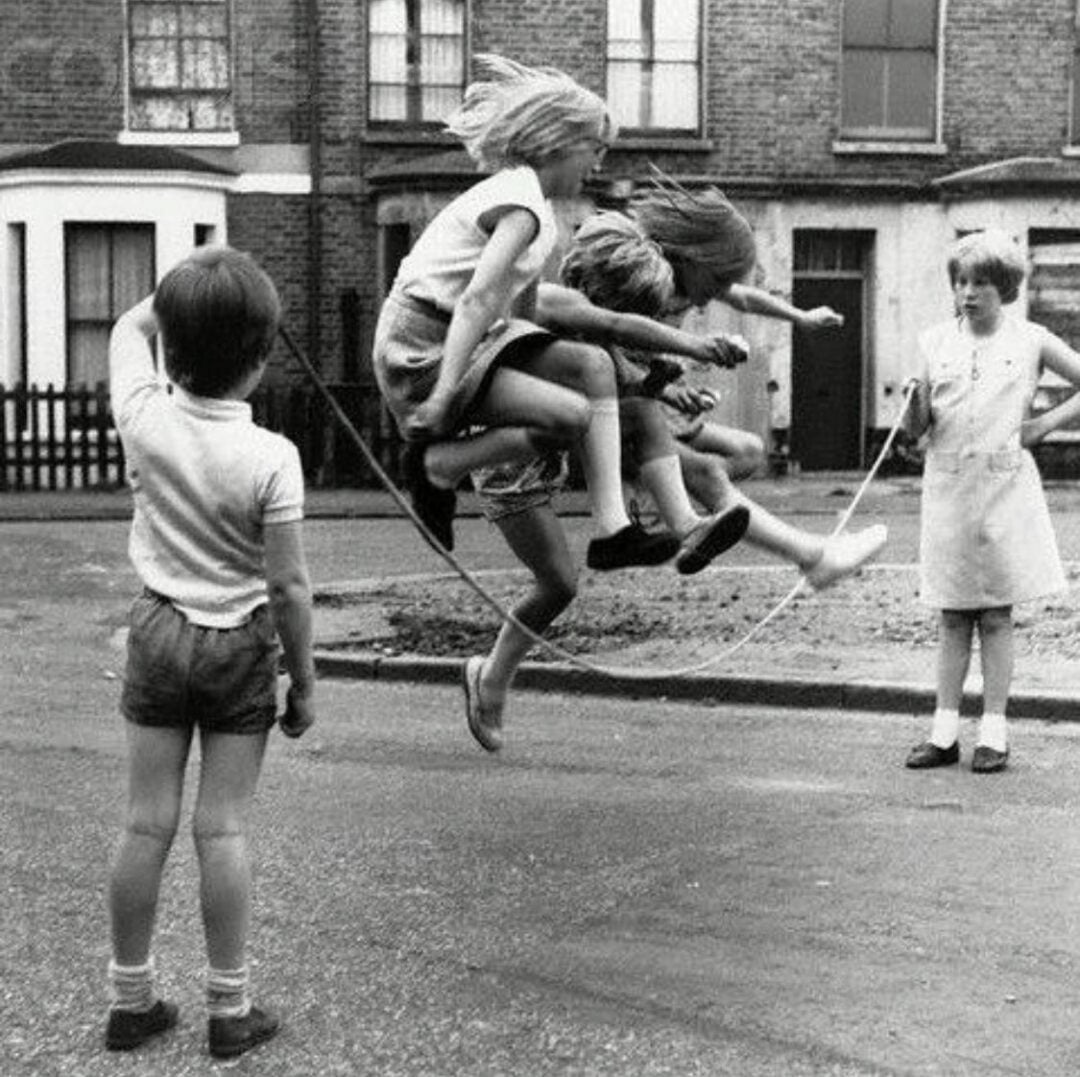 Four children are playing jump rope on a street. Three of them are jumping together while another child swings the rope. The background shows brick houses and a picket fence. The scene is in black and white.