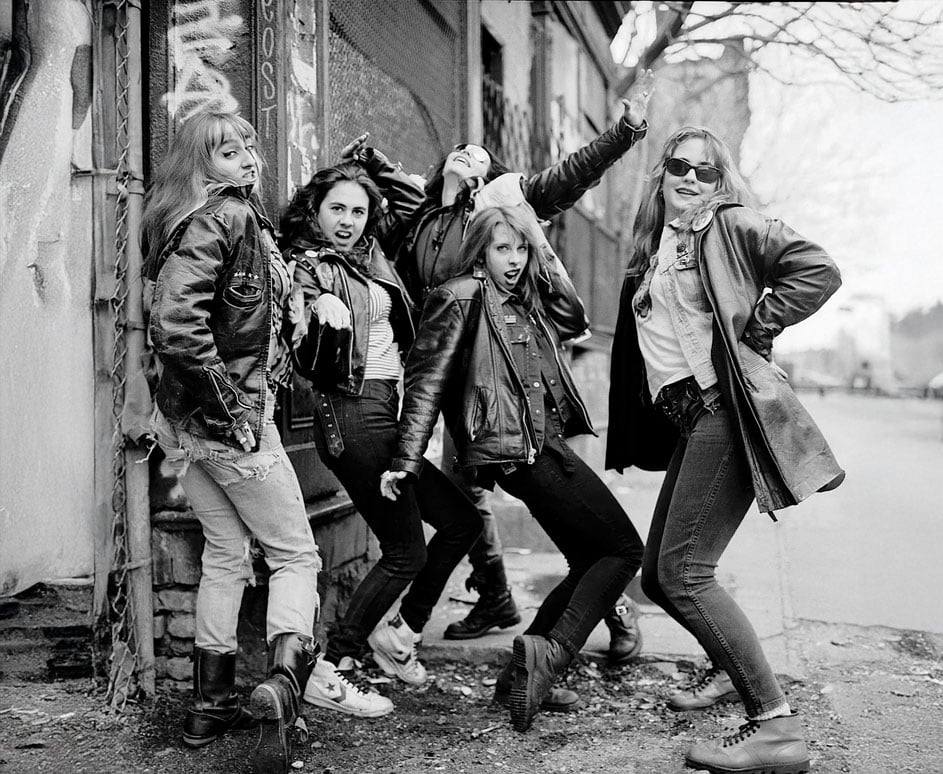 Five women pose energetically on a city sidewalk, wearing leather jackets, jeans, and boots. Graffiti and a brick wall are visible behind them, creating an edgy, urban atmosphere.