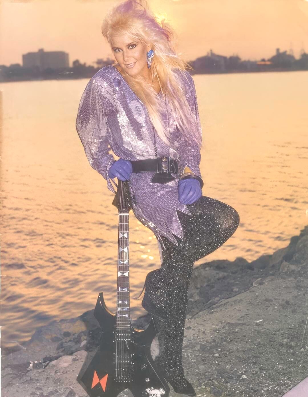 A woman with long blond hair, wearing a sparkly purple top, glittery black pants, blue gloves, and bold earrings, poses by the water at sunset with a black electric guitar. The name "Lita Ford" appears in the bottom left corner.