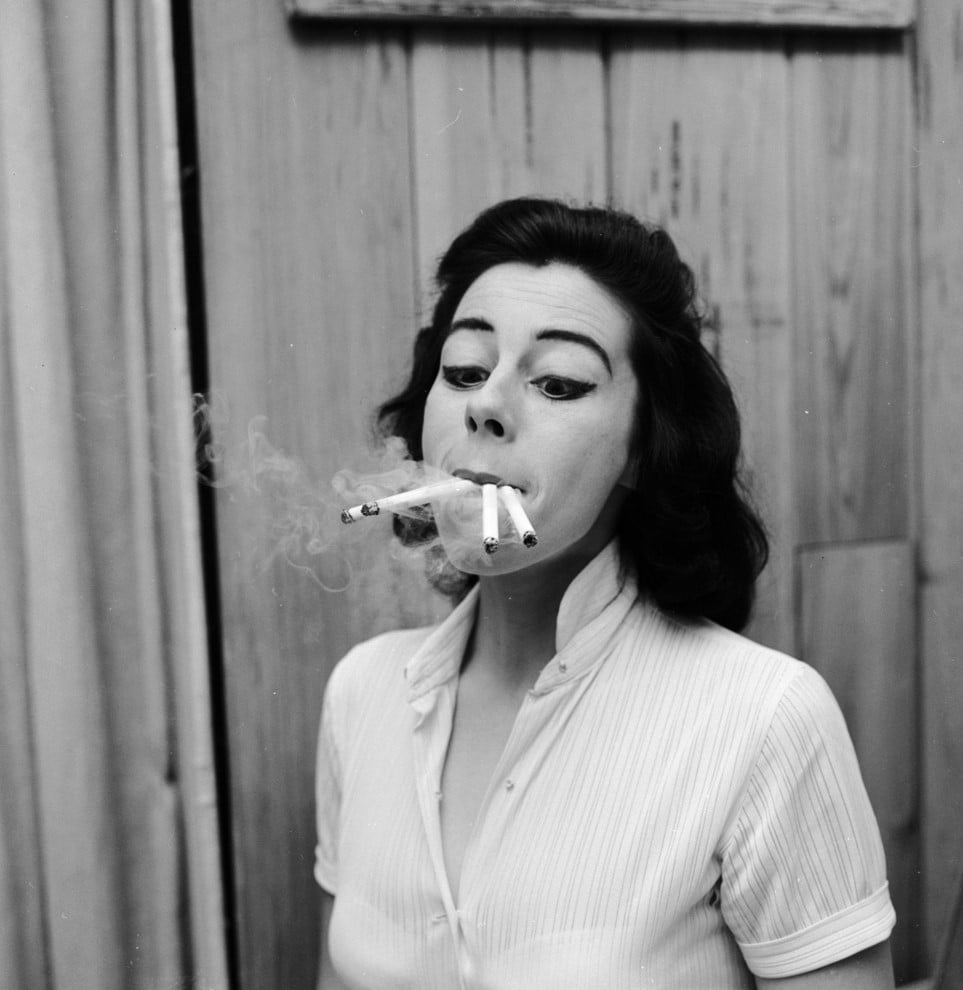 A woman with dark hair and dramatic eye makeup smokes three cigarettes at once, holding all three in her mouth, as smoke drifts upward. She stands in front of a wooden background, wearing a light-colored blouse.