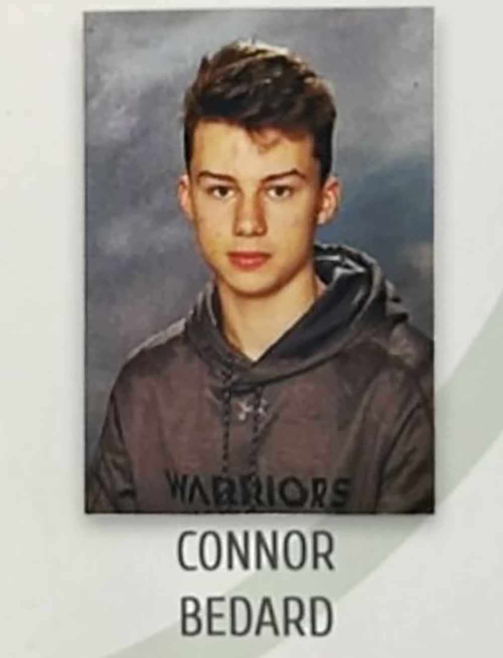A young person with short brown hair wearing a dark hoodie that says "WARRIORS" poses for a school portrait in front of a gray background. Text below the photo reads “Connor Bedard.”