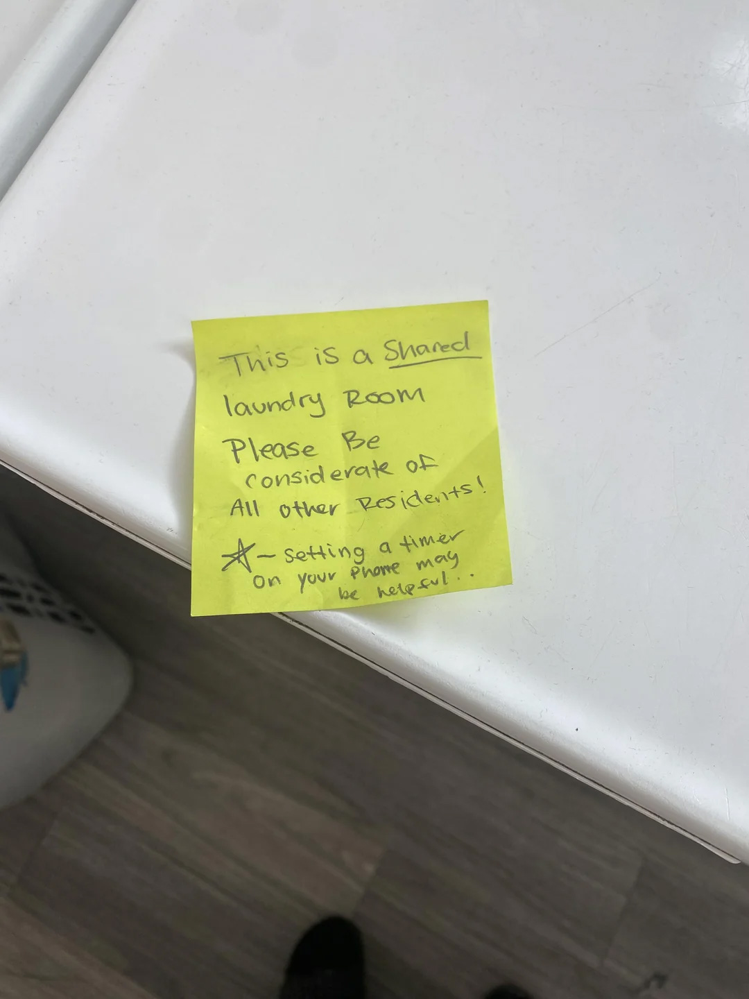 A yellow sticky note on a white surface reads: "This is a shared laundry room. Please be considerate of all other residents! —Setting a timer on your phone may be helpful.