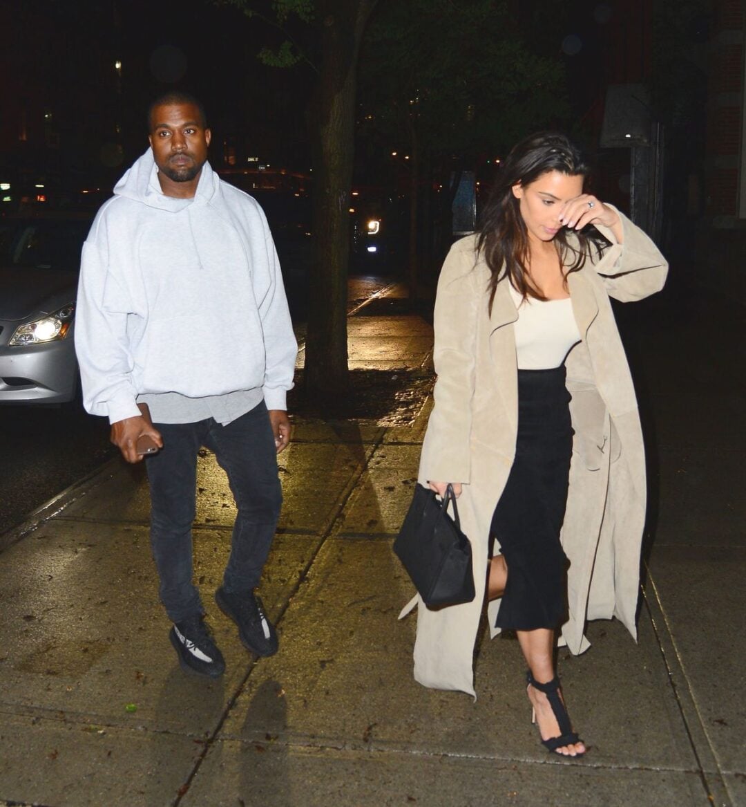 A man in a gray hoodie and black pants walks on a wet sidewalk at night beside a woman in a long beige coat, cream top, and black skirt. She holds a black handbag and touches her face as they walk.