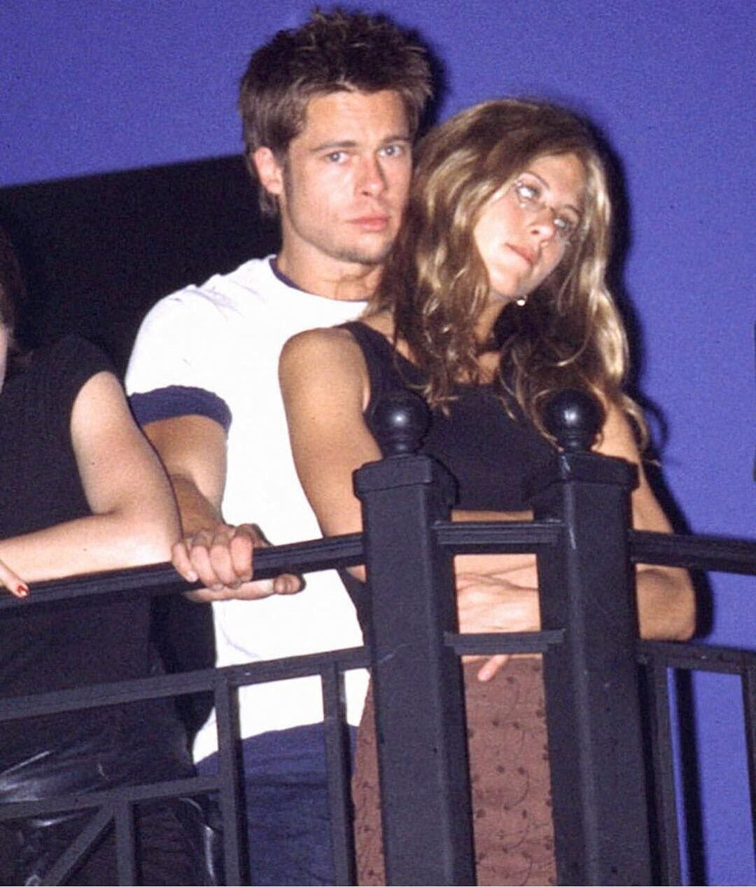 A young man in a white shirt stands closely behind a woman in a black top and brown skirt, both leaning on a black metal railing against a deep blue background. The woman looks to the side with her arms crossed.