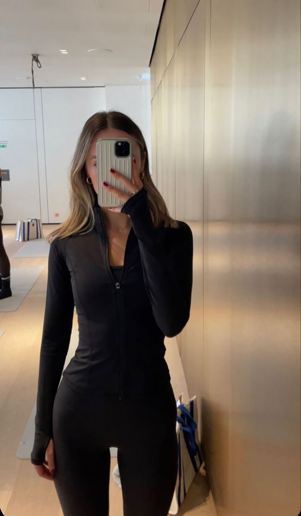 A woman in black athletic wear takes a mirror selfie in a modern, brightly lit room with light wood floors and gold-toned wall panels. Her phone covers part of her face.