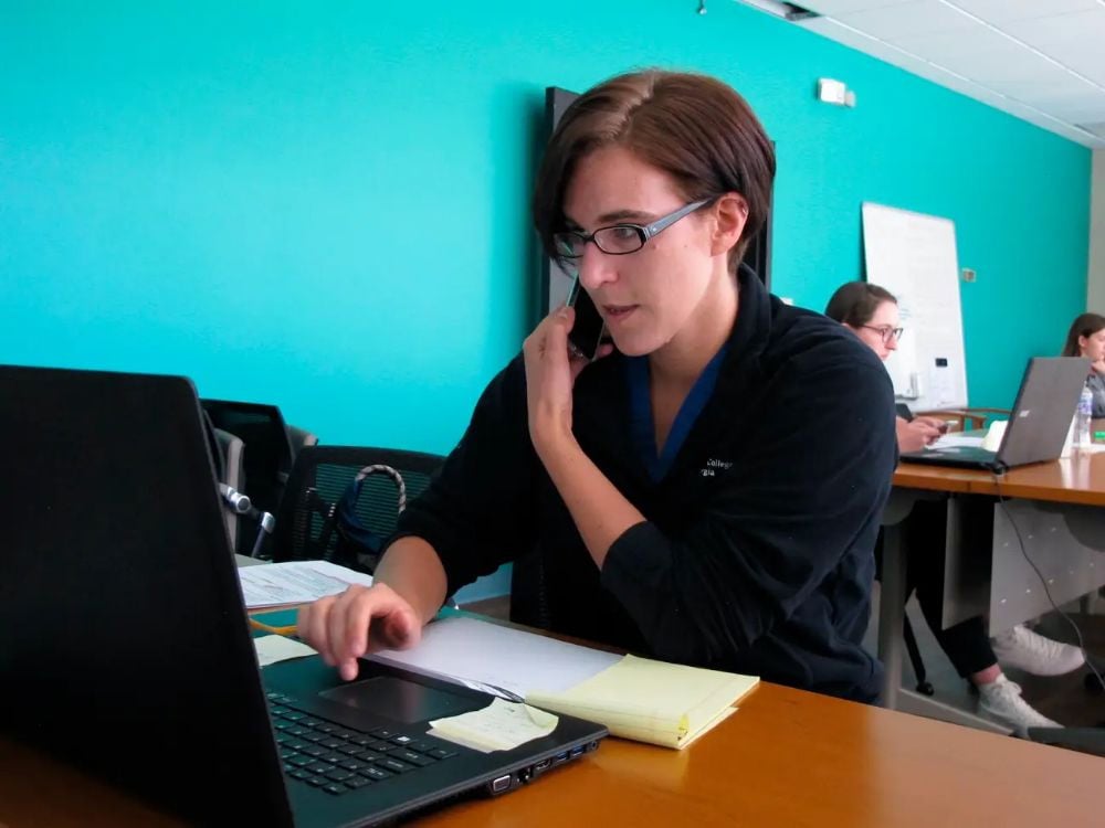 A woman wearing glasses sits at a desk using a laptop and talking on the phone, with a notepad in front of her. Other people work at desks in the background in a brightly lit room with teal walls.