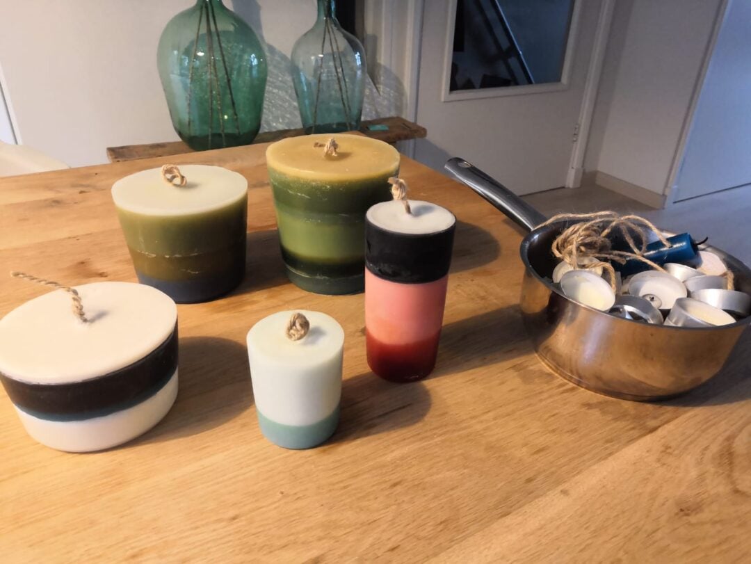 Five colorful, handmade candles in various sizes are arranged on a wooden table next to a pot filled with used tealight holders and candle wicks. Green glass vases and a mirror are in the background.