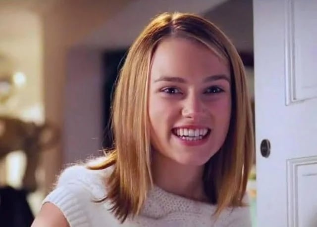 A young woman with straight blonde hair and a white sweater smiles brightly while standing by a door indoors.