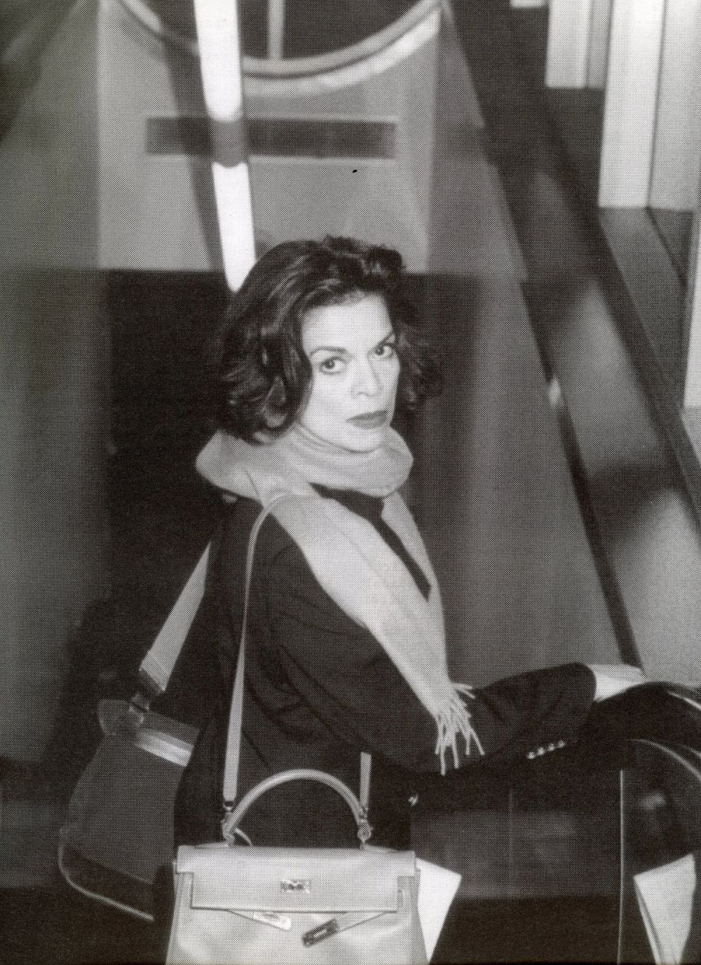 A person with shoulder-length hair and a scarf stands on an escalator, looking back over their shoulder with a serious expression. They are carrying a handbag and wearing a dark coat. The image is in black and white.