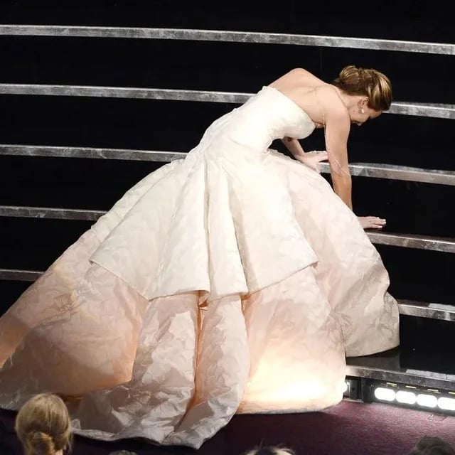 A woman in a strapless, white ball gown is stumbling and reaching out to support herself as she climbs stairs at a formal event.