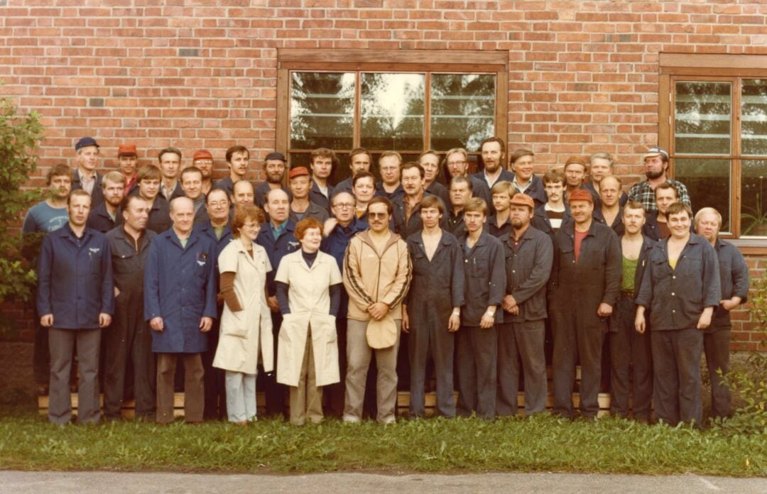 A large group of men and two women, mostly dressed in work clothes and overalls, stand in rows outside in front of a brick building with windows, posing for a group photo.